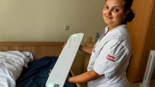 Een zorgmedewerker praat met een bewoonster die in bed ligt, terwijl ze de app van de bedsensor checkt.