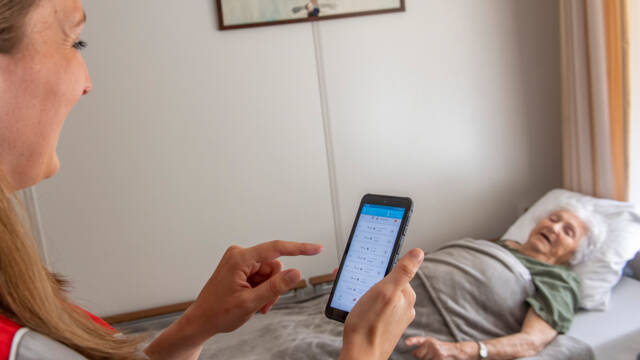 Een zorgmedewerker praat met een bewoonster die in bed ligt, terwijl ze de app van de bedsensor checkt.