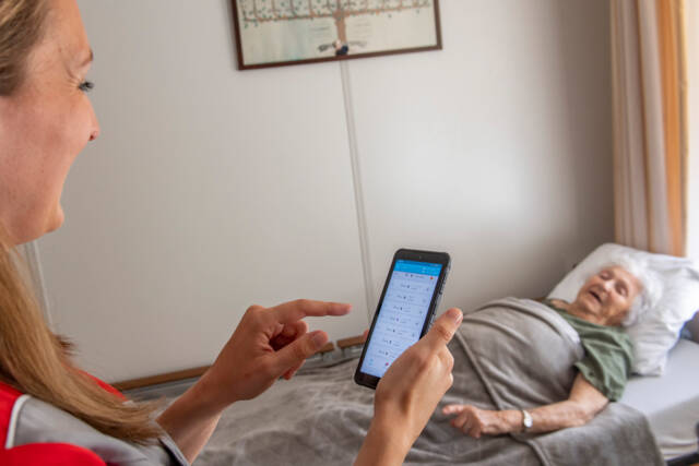 Een zorgmedewerker praat met een bewoonster die in bed ligt, terwijl ze de app van de bedsensor checkt.